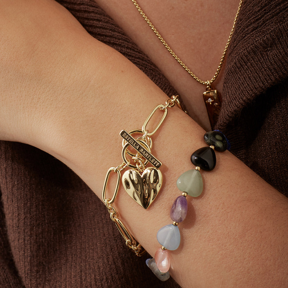 Gold bracelet with heart charm and multicolored beads on a wrist against a brown background