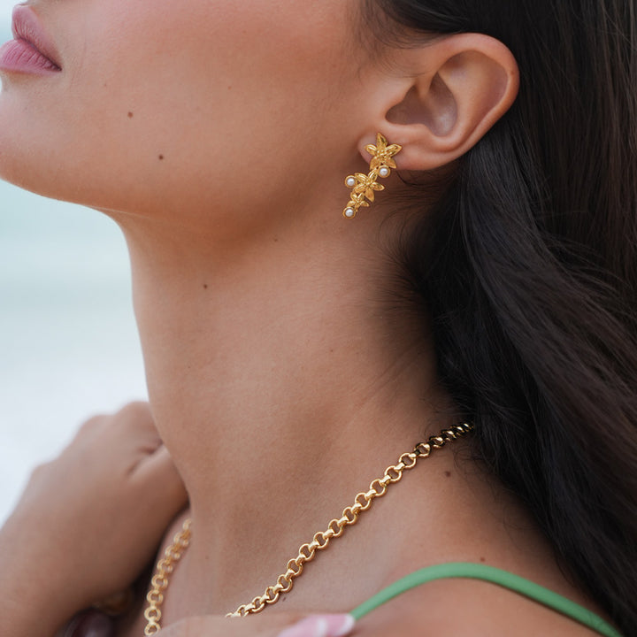 woman wearing gold drop earrings with 3 flowers and 3 small pearls and a gold chain necklace