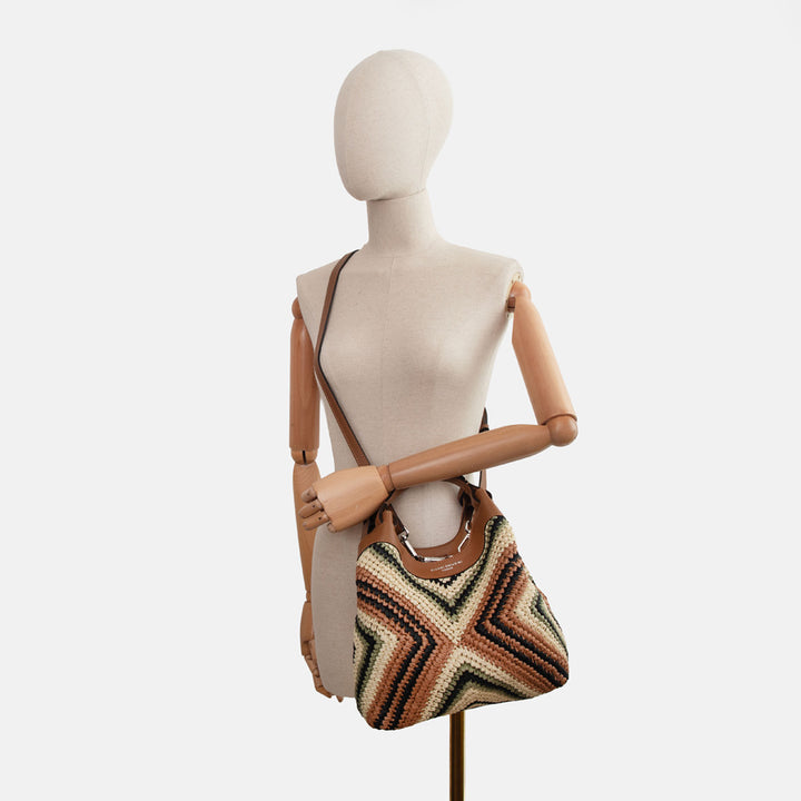 Mannequin wearing a brown, beige and green chevron design woven straw handbag with brown leather handles and crossbody strap