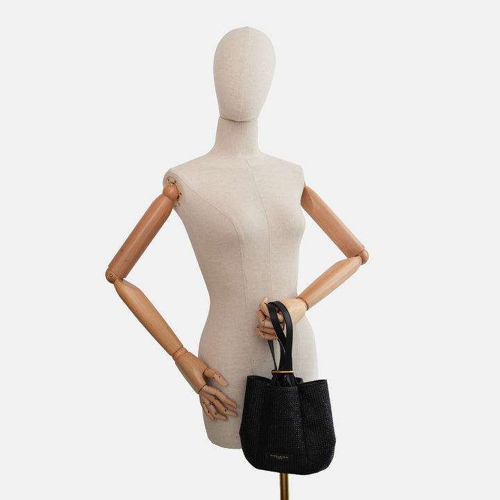 Mannequin holding a black woven straw bucket handbag with black leather straps and gold ring closure on a white background