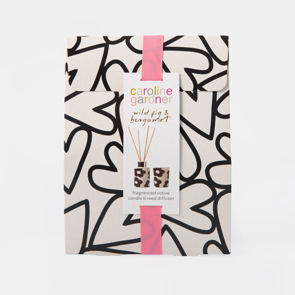 A packaged reed diffuser set from Caroline Gardner, featuring a white box with a black abstract hearts pattern and a pink ribbon. The front has a white paper label with the brand name and product name 'wild fig & bergamot fragranced votive candle & reed diffuser'.