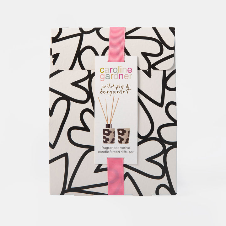 A packaged reed diffuser set from Caroline Gardner, featuring a white box with a black abstract hearts pattern and a pink ribbon. The front has a white paper label with the brand name and product name 'wild fig & bergamot fragranced votive candle & reed diffuser'.