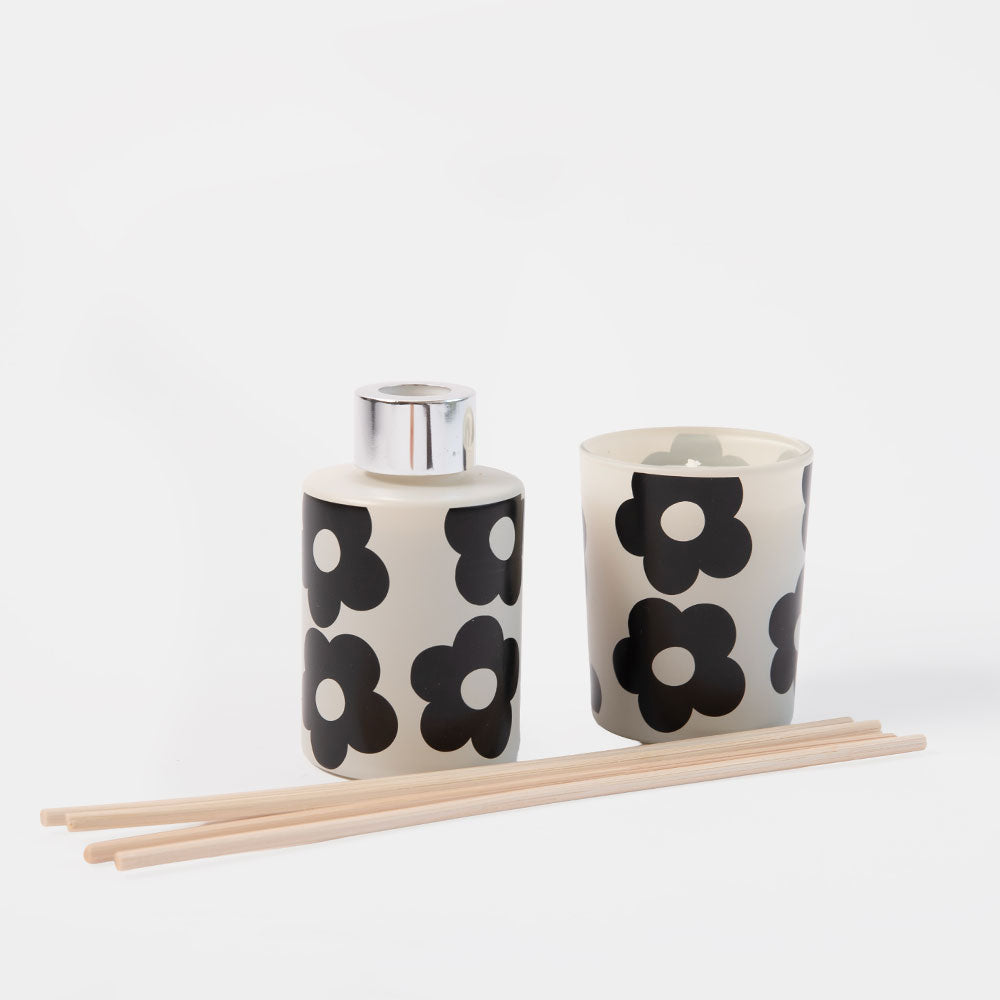 A ceramic diffuser and candle set with a black and white floral pattern, accompanied by four natural wood reeds, on a clean white background.