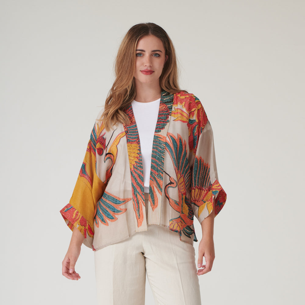 woman wearing a short kimono in orange, green & blue cranes print on a stone base with a white t-shirt & cream trousers
