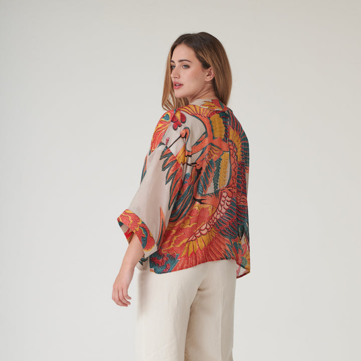 woman wearing a short kimono in orange, green & blue cranes print on a stone base with cream trousers