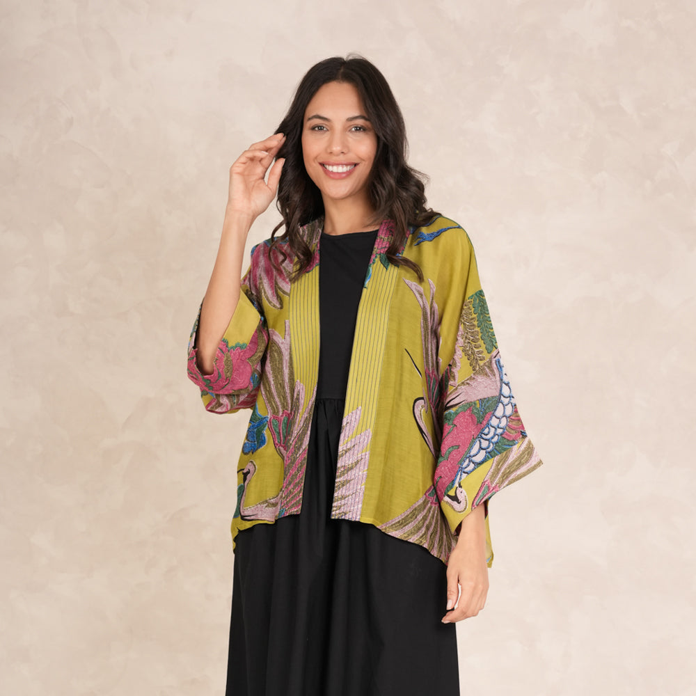 woman wearing a short kimono with pink, green & blue cranes design on an olive base over a black dress