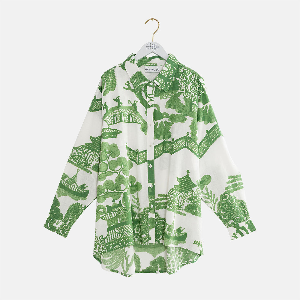 A long-sleeve button-up shirt with a green and white scenic print, hanging on a white hanger against a light gray background.