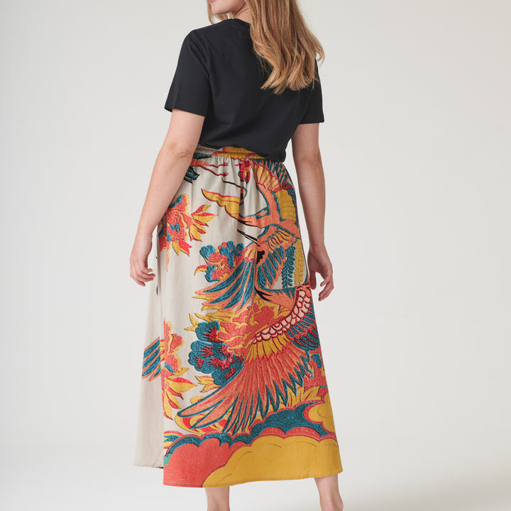 woman wearing an a-line skirt with design of cranes in orange, green & blue on a stone base with a black t-shirt