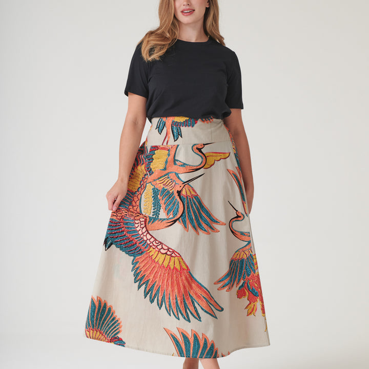 woman wearing an a-line skirt with design of cranes in orange, green & blue on a stone base with a black t-shirt