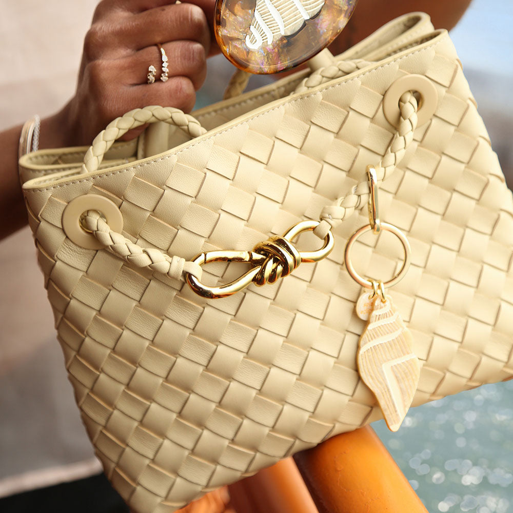 Beige woven handbag with gold accents held by a person with a seashell bag charm