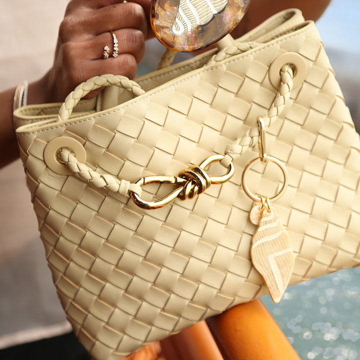 Beige woven handbag with gold accents held by a person with a seashell bag charm