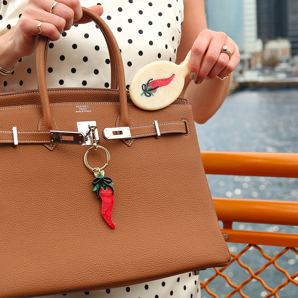 Brown handbag with a red chili pepper charm held by a person in a polka dot dress, with a blurred waterfront background.