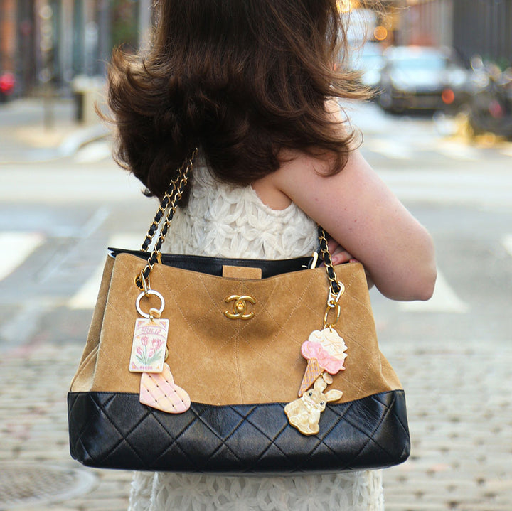 Person holding a tan and black handbag with decorative charms on a city street.