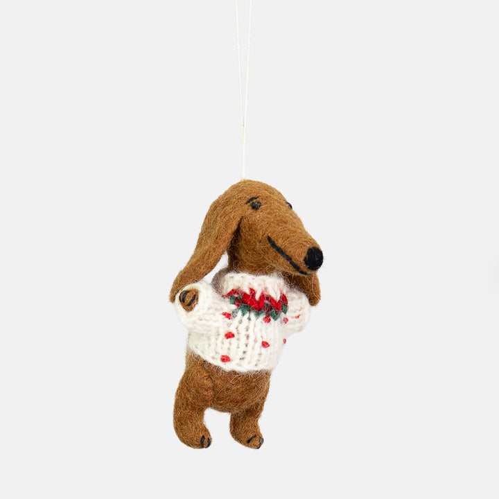 A felt sausage dog hanging ornament wearing a white knit sweater with red and green holiday accents, suspended by a thin white string against a light gray background.