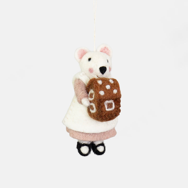 A felted wool mouse ornament wearing a white dress and pink sleeves, holding a brown gingerbread house with white polka dots, hanging against a plain light gray background.