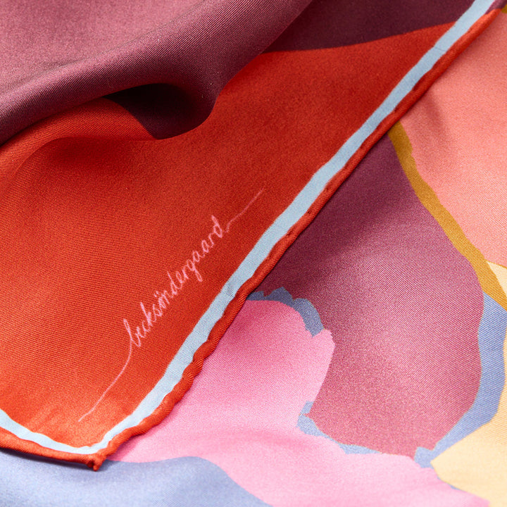 A close-up of a folded silk scarf with a vibrant abstract pattern in orange, pink, purple, and yellow, featuring a white border and the brand name 'lindberghaard' printed in white script.