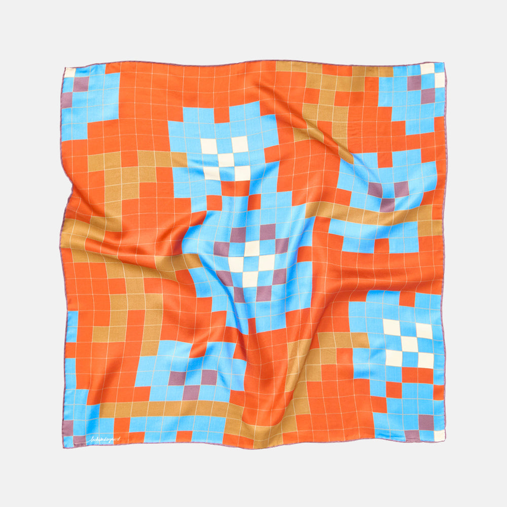 A square silk scarf with a colorful geometric pixelated pattern, laid flat on a light gray background. The pattern features orange, light blue, tan, white, and purple blocks.