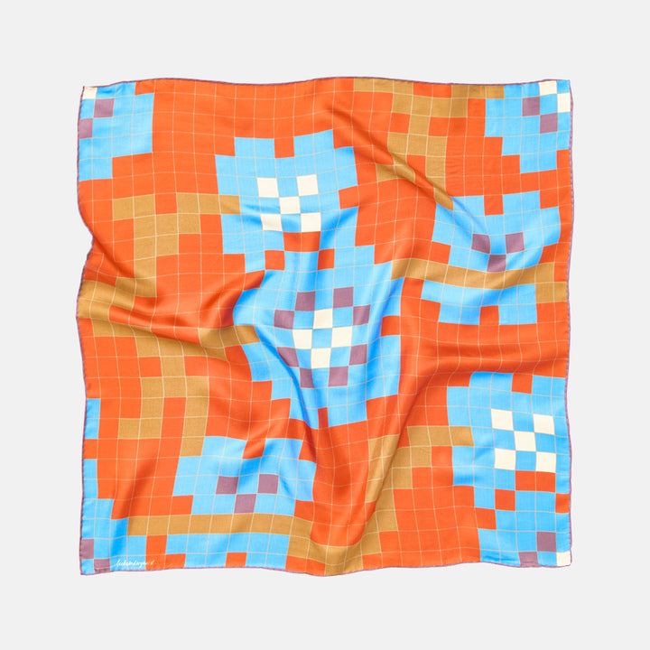 A square silk scarf with a colorful geometric pixelated pattern, laid flat on a light gray background. The pattern features orange, light blue, tan, white, and purple blocks.