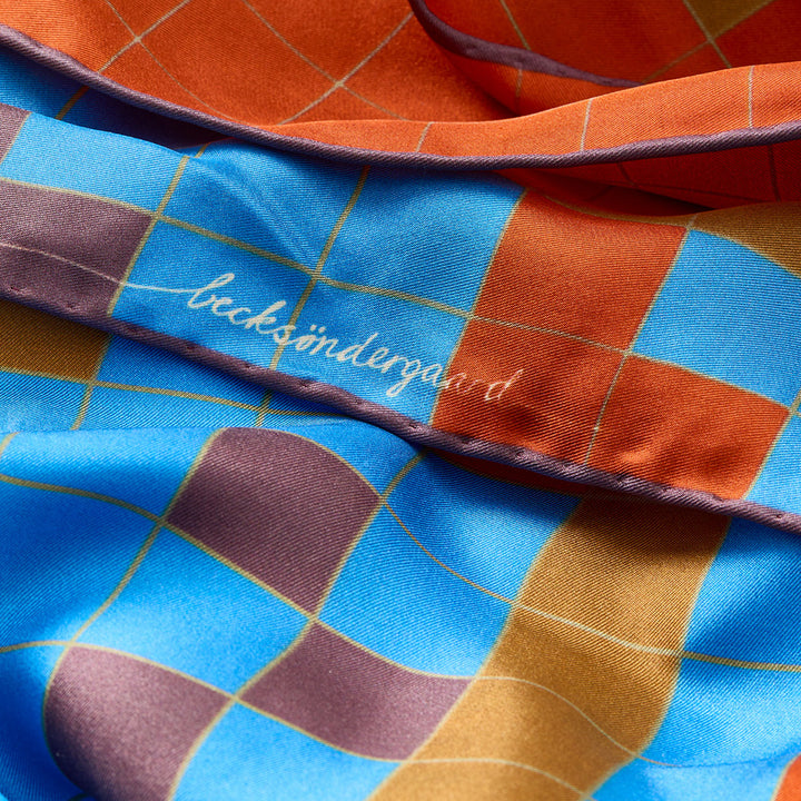 A close-up of a silk scarf with a checkered pattern in blue, orange, and purple, featuring gold grid lines and the brand name 'becksøndergaard' embroidered in white script.