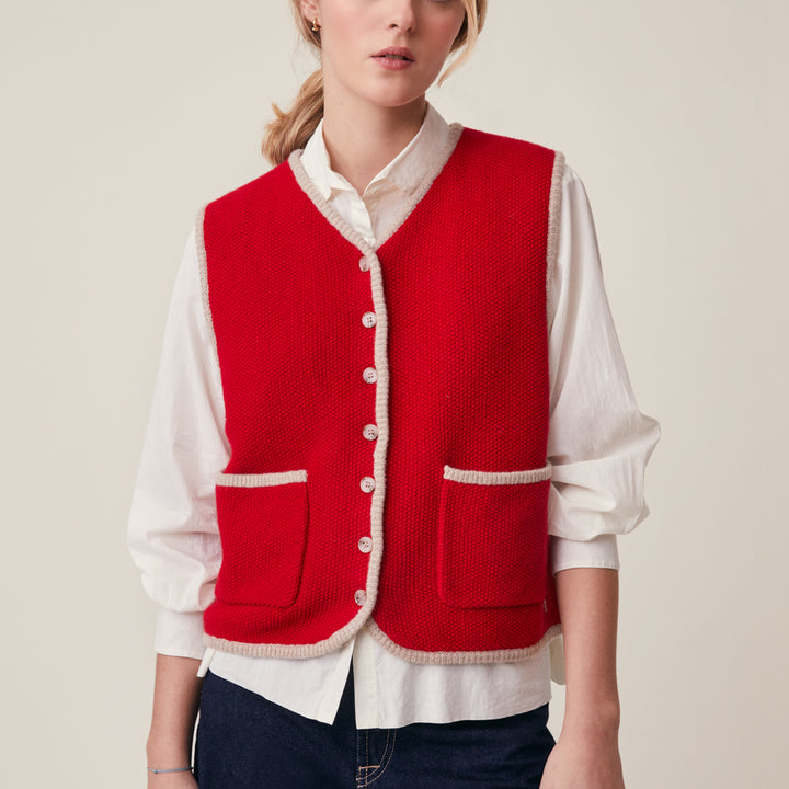 A woman wearing a bright red knit vest with cream trim over a white button-up shirt and dark blue jeans, photographed against a plain light beige studio background.