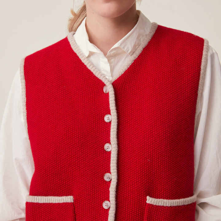 A close-up of a person wearing a bright red knit vest over a white collared shirt. The vest has cream-colored trim along the neckline, armholes, and pocket edges, with matching cream buttons down the front.