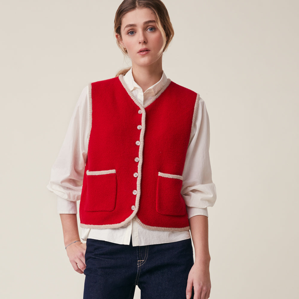 A woman modeling a bright red textured knit vest with cream trim over a cream button-up shirt and dark blue jeans, set against a solid light beige background.