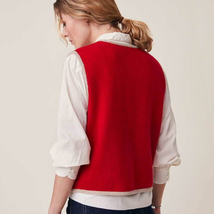 A woman shown from the back wearing a bright red sleeveless knit vest over a white long-sleeve blouse, against a plain light beige studio background.