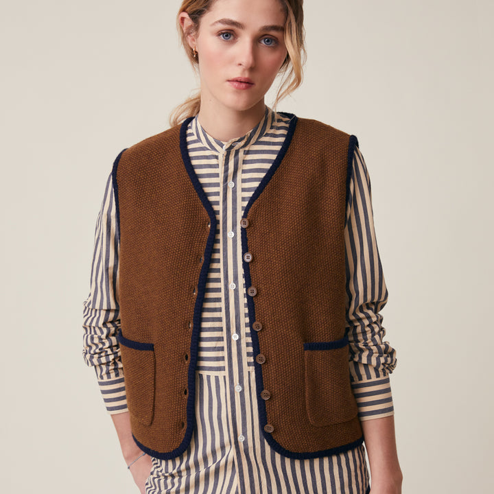 A woman wearing a brown knit button-front vest with dark navy trim over a cream and navy striped shirt, photographed against a plain light beige studio background.