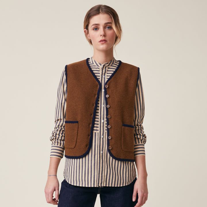 A woman modeling a brown knitted vest with navy blue trim over a beige and navy striped button-down shirt, paired with dark jeans, against a solid light beige background.
