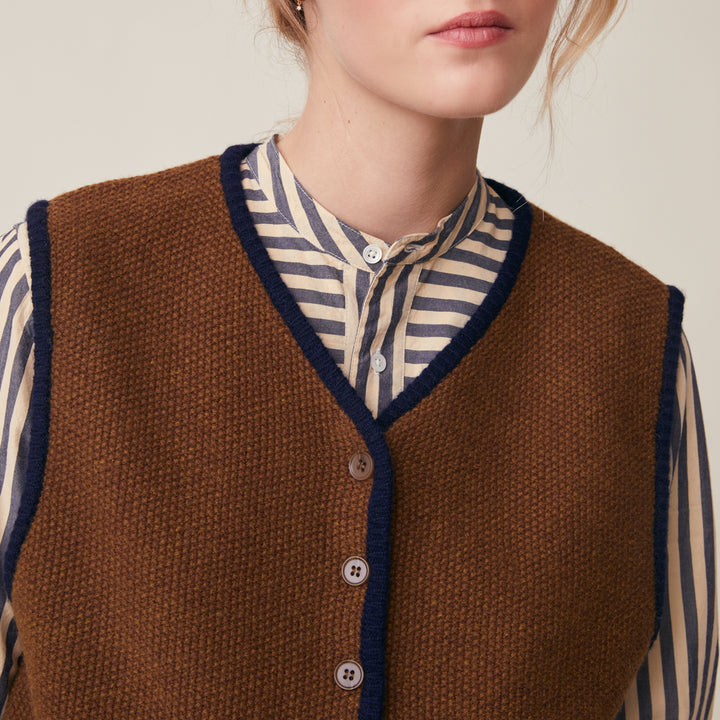A close-up of a person wearing a brown knit vest with dark navy trim over a cream and navy striped button-up shirt, against a plain light beige background.