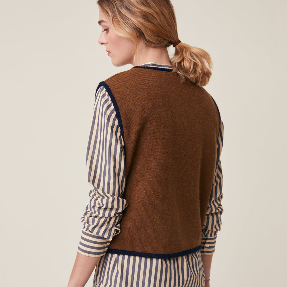A woman shown from the back wearing a brown knit sweater vest over a cream and navy blue striped shirt, set against a plain light beige studio background.