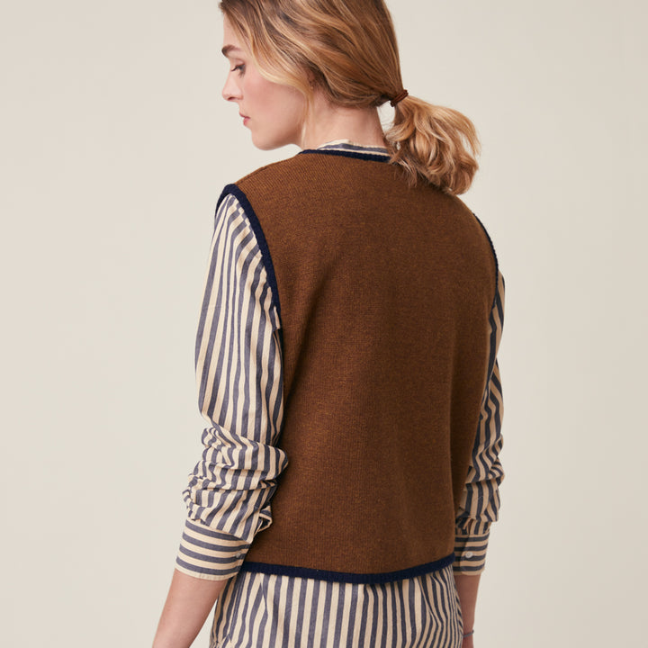 A woman shown from the back wearing a brown knit sweater vest over a cream and navy blue striped shirt, set against a plain light beige studio background.