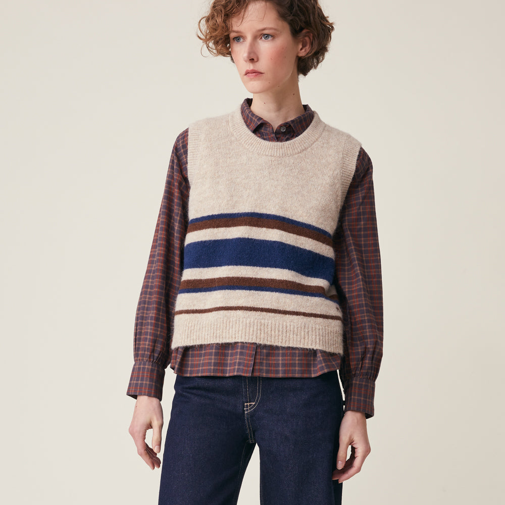A model wearing a cream-colored knit sweater vest with horizontal navy, brown, and beige stripes across the chest, layered over a brown and red plaid button-down shirt and paired with dark blue jeans, set against a plain beige background.