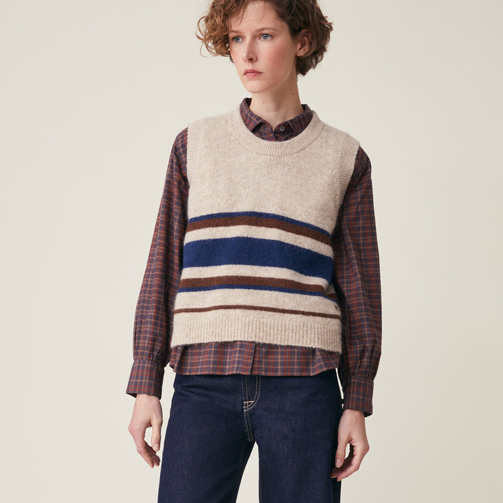 A model wearing a cream-colored knit sweater vest with horizontal navy, brown, and beige stripes across the chest, layered over a brown and red plaid button-down shirt and paired with dark blue jeans, set against a plain beige background.