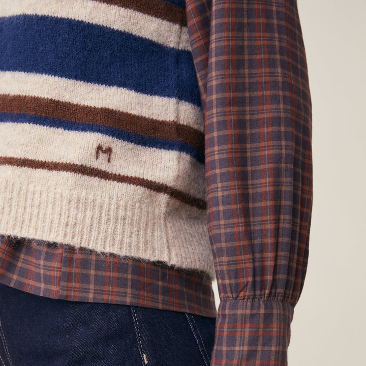 Close-up of a striped knit sweater with cream, navy, and brown horizontal bands, layered over a long-sleeve plaid shirt in navy, red, and orange tones, paired with dark blue jeans.