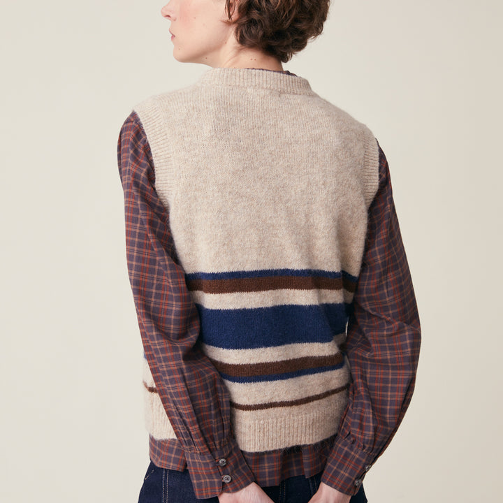 A person shown from the back wearing a knit sweater vest layered over a long-sleeve plaid button-up shirt. The vest is cream with horizontal navy, brown, and cream stripes near the hem, and the sleeves/shirt portion are in a muted red-brown plaid.