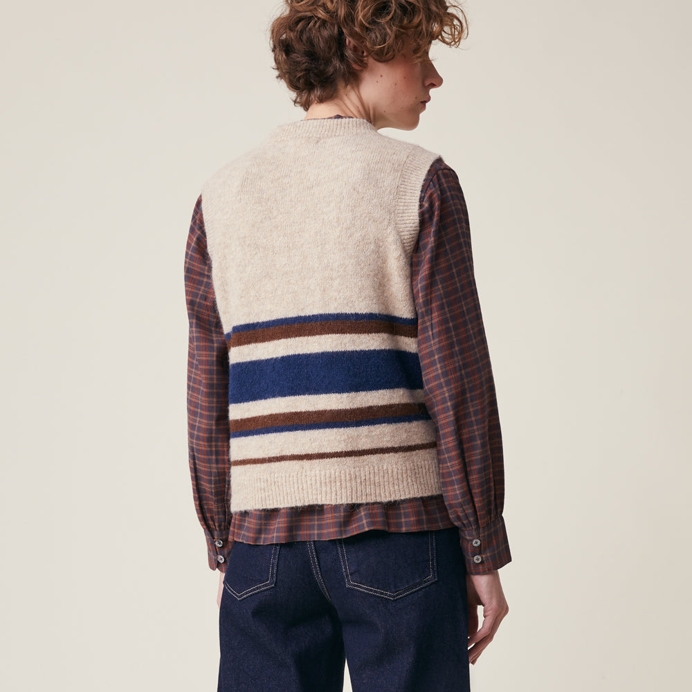A model shown from the back wearing a cream knit sweater vest layered over a red and brown plaid flannel shirt, paired with dark blue jeans, against a solid light beige background.