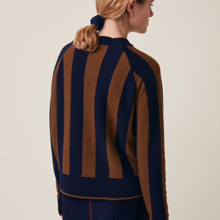 A woman shown from the back wearing a knit sweater with bold vertical stripes in navy blue and brown, paired with matching navy bottoms, against a plain light beige studio background.