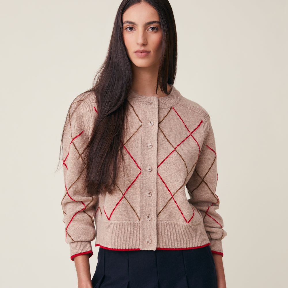 A woman wearing a beige knit cardigan with an argyle pattern in red and brown, paired with dark navy trousers, against a solid light beige background.