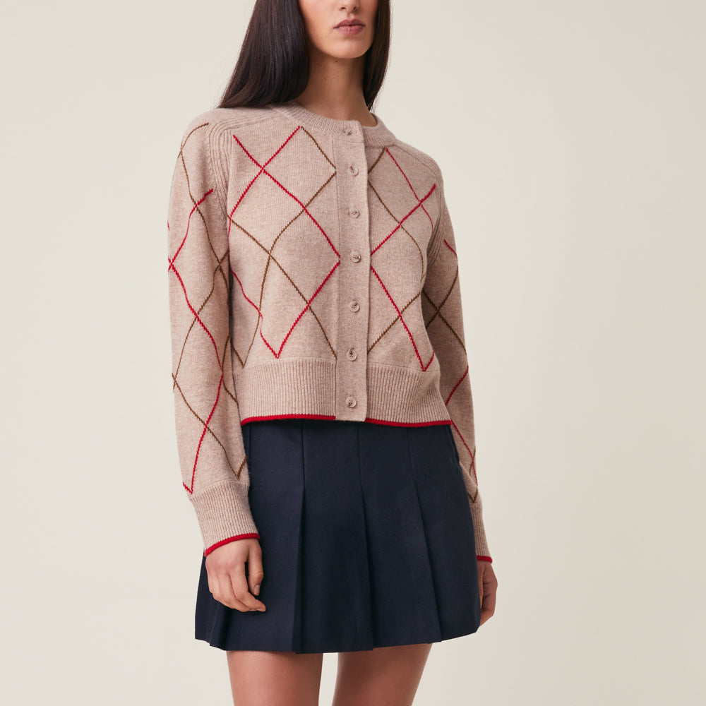A woman models a beige knit cardigan with an argyle pattern in red and brown, paired with a dark navy pleated skirt, against a solid light beige background.