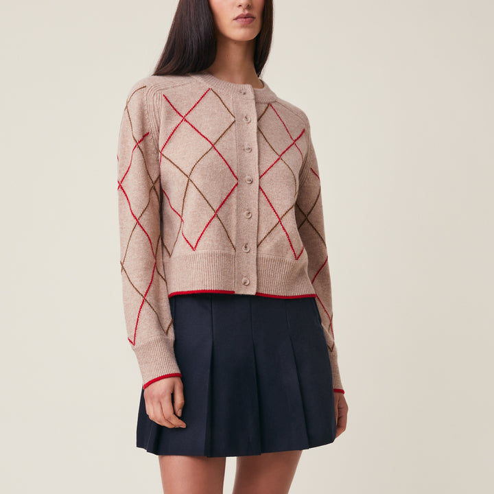 A woman models a beige knit cardigan with an argyle pattern in red and brown, paired with a dark navy pleated skirt, against a solid light beige background.