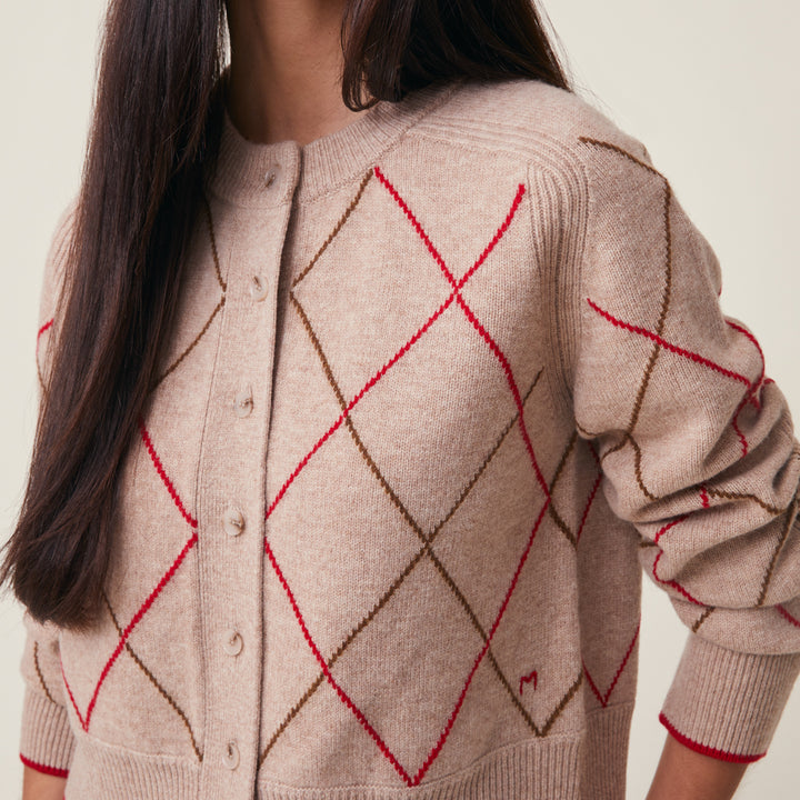 A close-up of a person wearing a beige knit cardigan with an argyle pattern in brown and red, featuring a button-up front and ribbed cuffs and hem.