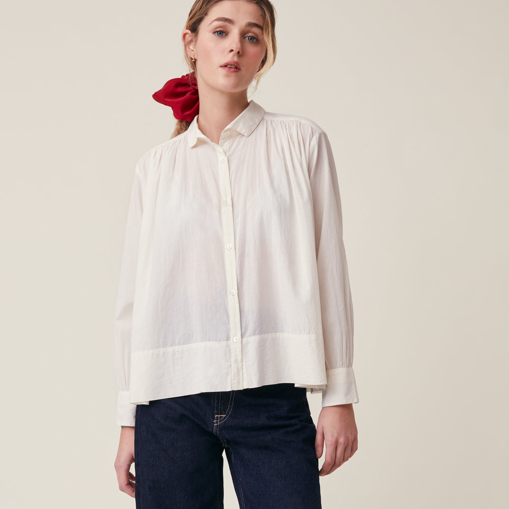 A woman wearing a white long-sleeve button-up blouse with gathered detailing at the shoulders and hem, paired with dark blue jeans and a red scrunchie, against a plain beige studio background.