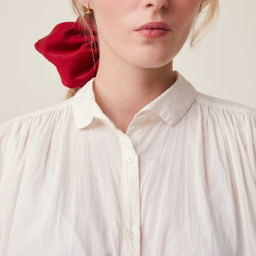 Close-up of a woman wearing a cream-colored, crinkled button-down blouse with a collar and pleated shoulders, styled with a bright red fabric hair scrunchie.