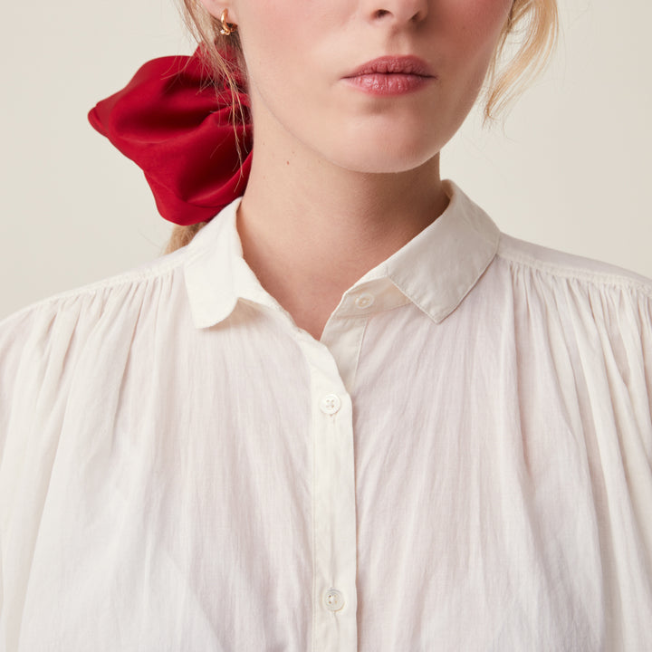 Close-up of a woman wearing a cream-colored, crinkled button-down blouse with a collar and pleated shoulders, styled with a bright red fabric hair scrunchie.