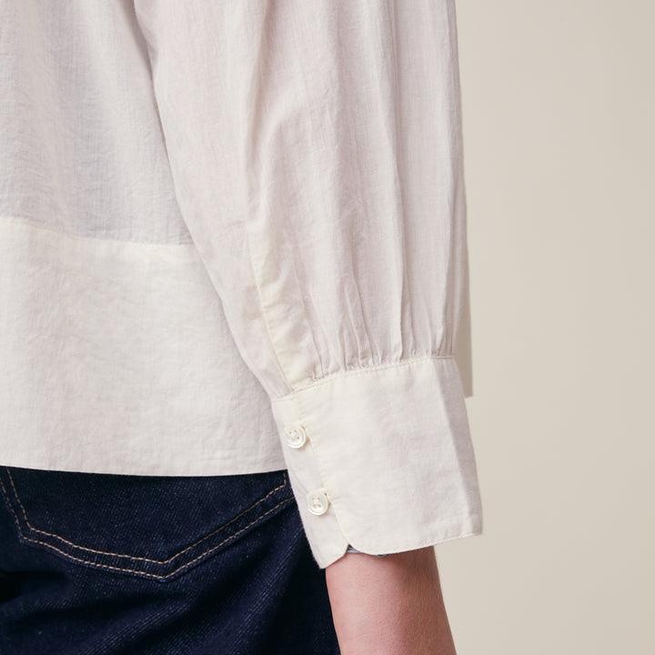 Close-up of a person wearing a light cream or off-white long-sleeve blouse with a gathered cuff and three buttons, paired with dark blue jeans against a plain light beige background.