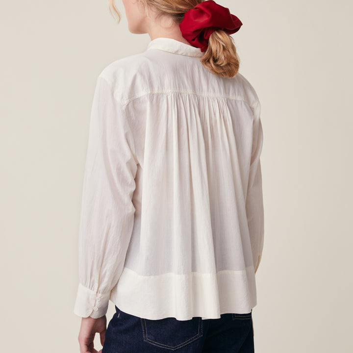 A woman shown from the back wearing a white long-sleeve blouse with a gathered, smocked back panel and a pleated neckline, paired with dark blue jeans and a red hair scrunchie, against a light beige background.