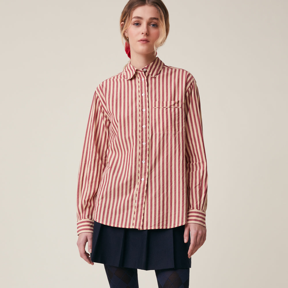 A woman modeling a long-sleeve button-up shirt with vertical red and beige stripes, paired with a dark navy pleated skirt, against a plain light beige studio background.