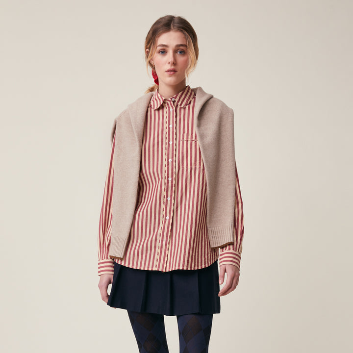 A model stands against a plain beige studio background wearing a beige knit hooded cardigan over a red and white vertically striped button-up shirt, paired with a dark pleated skirt and dark patterned tights.