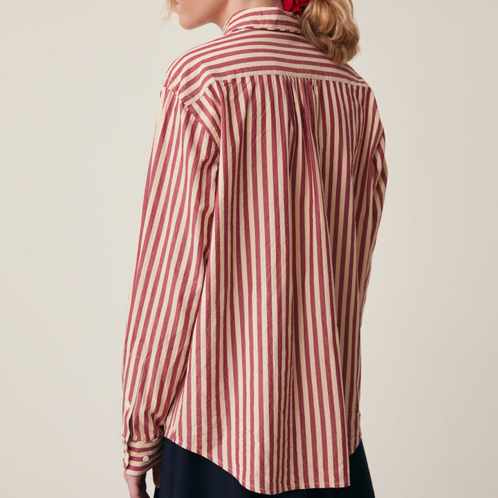 A woman shown from the back wearing a long-sleeve button-up shirt with vertical red and beige stripes, paired with a dark skirt. The photo is taken against a plain, light beige studio background.